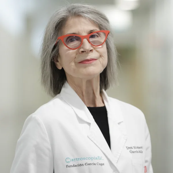 Portrait of Dr. Montserrat García Balletbó, specialist in regenerative medicine and co-founder of Instituto Cugat, wearing a white lab coat and red glasses in the clinic hallway.