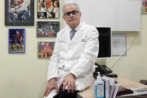 Dr Ramón Cugat sitting in his office wearing a white medical coat, surrounded by sports photos, representing his expertise in orthopaedic surgery and his work with elite football players recovering from ACL injuries.