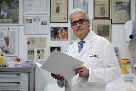 Dr. Ramon Cugat, in a white lab coat, stands in his office holding a folder. Sports memorabilia and articles adorn the wall behind him.